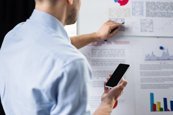 Cropped view of smartphone in hand of businessman working with flipchart on blurred background — Stock Photo