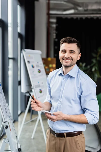 Hombre de negocios sonriente con lápiz y teléfono inteligente de pie cerca del rotafolio en la oficina - foto de stock