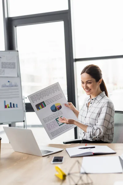 Lächelnde Geschäftsfrau zeigt bei Videoanruf auf Laptop auf Dokument mit Diagrammen — Stockfoto