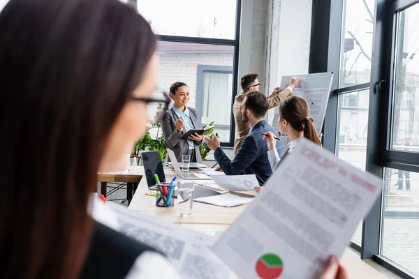 Interrassische Geschäftsleute, die mit Flipchart, Papieren und Laptops in der Nähe verschwommener Kollegen arbeiten — Stockfoto