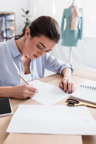 Näherin zeichnet Skizze nahe Smartphone und Schere auf Tisch — Stockfoto