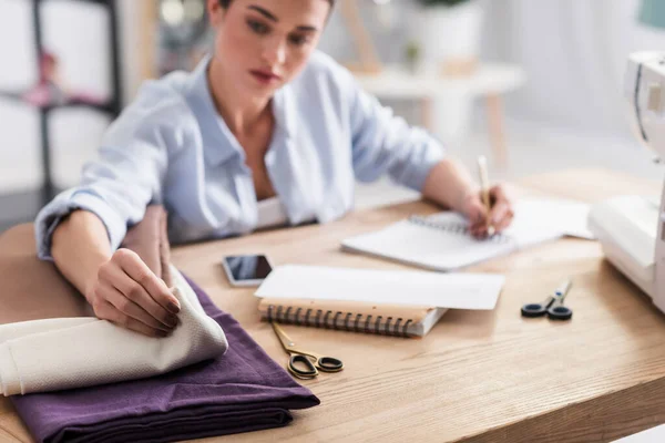 Unscharfe Näherin nimmt Tuch und schreibt auf Notizbuch in der Nähe des Smartphones — Stockfoto