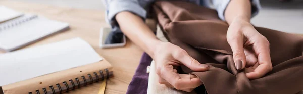 Vue recadrée du tissu de maintien de couturière près du téléphone cellulaire flou et des ordinateurs portables, bannière — Photo de stock