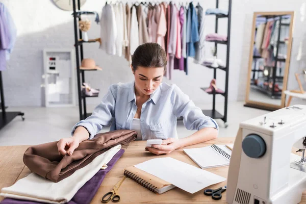 Costurera usando teléfono inteligente cerca de tela, cuadernos y máquina de coser - foto de stock