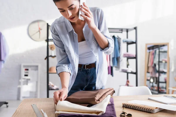 Costurera hablando en el teléfono inteligente cerca de tela y cuadernos en la mesa - foto de stock