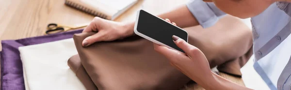 Cropped view of seamstress using smartphone near blurred fabric, banner — Stock Photo