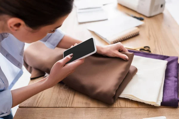 Costurera borrosa con teléfono celular sosteniendo tela cerca de tijeras en la mesa - foto de stock