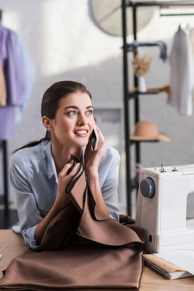 Cheerful seamstress talking on smartphone near notebook and sewing machine on table — Stock Photo