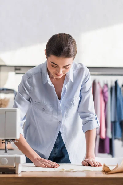 Joven costurera de pie cerca de tela y máquina de coser borrosa en el estudio — Stock Photo