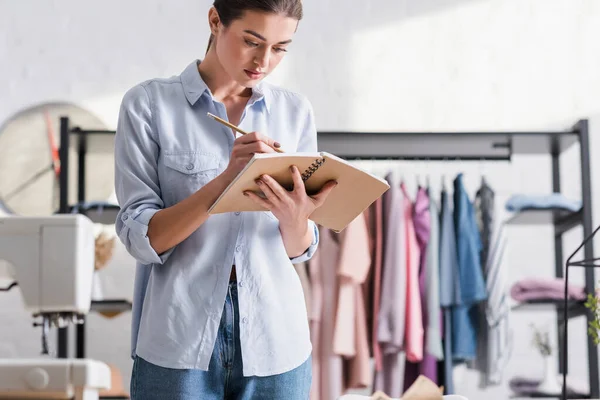 Seamstress writing on notebook near blurred sewing machine — Stock Photo