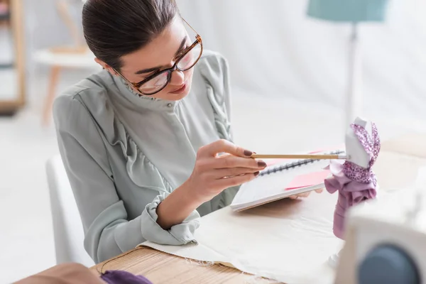 Seamstress with pencil and notebook looking at mannequin with dress on table — Stock Photo