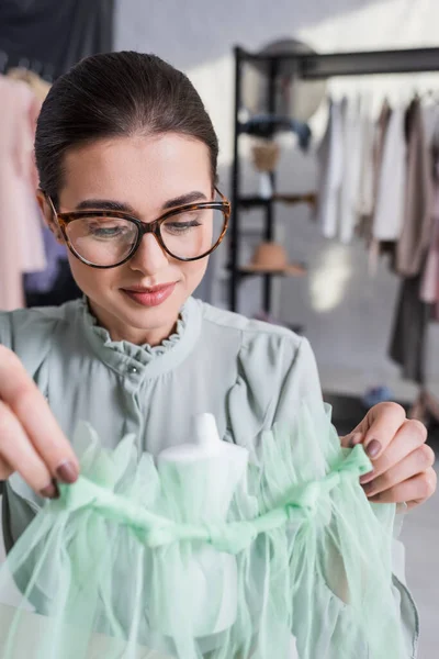 Costurera en gafas que sostiene la tela cerca del maniquí borroso - foto de stock