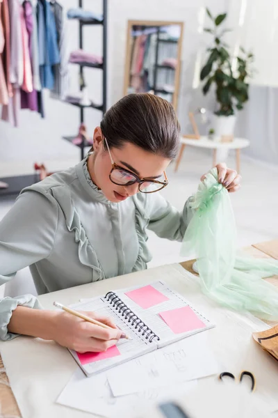 Seamstress holding cloth and writing on notebook near sketches on table — Stock Photo