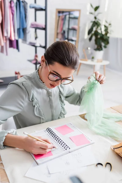 Seamstress segurando pano e escrevendo no caderno perto de esboços na mesa — Fotografia de Stock