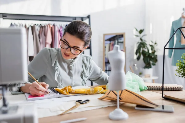 Diseñador escribiendo en el cuaderno cerca de tela, teléfono inteligente y maniquí en la mesa - foto de stock