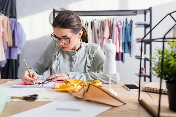 Diseñador escribiendo en el portátil cerca de teléfono inteligente y tela en el estudio - foto de stock