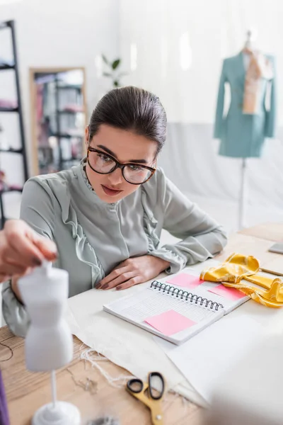 Seamstress segurando manequim perto notebook e tesoura borrada na mesa — Fotografia de Stock