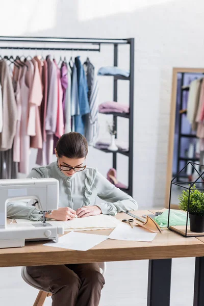 Tissu de marquage designer tout en travaillant près de croquis et machine à coudre en studio — Photo de stock