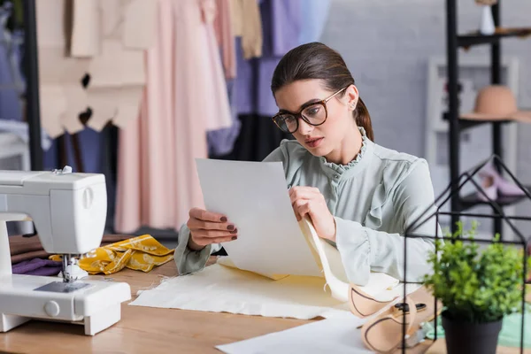 Costurera sosteniendo papel cerca de la tela y máquina de coser en la mesa - foto de stock