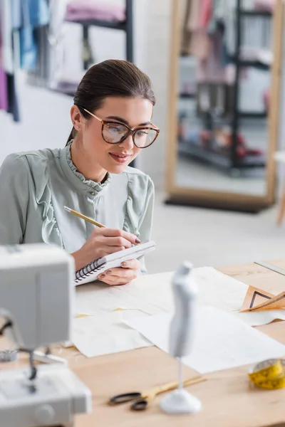 Couturière souriante écrivant sur le cahier près du mannequin et du tissu sur la table — Photo de stock