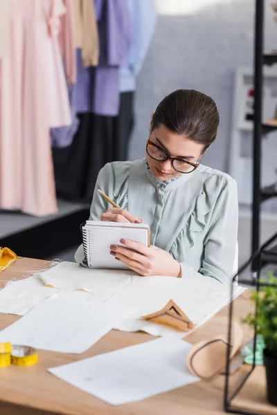 Designer writing on notebook near fabric and sewing patterns on table — Stock Photo