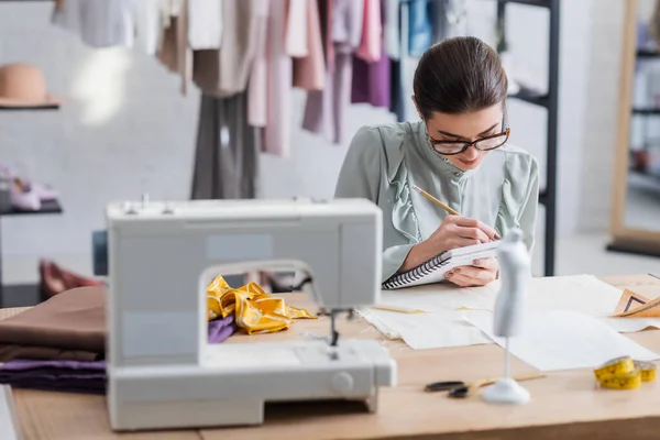 Näherin schreibt auf Notizbuch neben Stoff und verschwommener Nähmaschine — Stockfoto