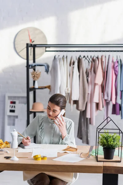 Seamstress falando no smartphone perto de tecido e notebook — Fotografia de Stock