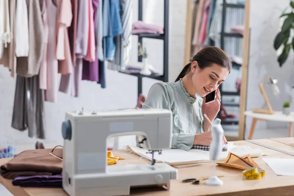 Designer parler sur smartphone et écrire sur ordinateur portable près du tissu et de l'équipement — Photo de stock