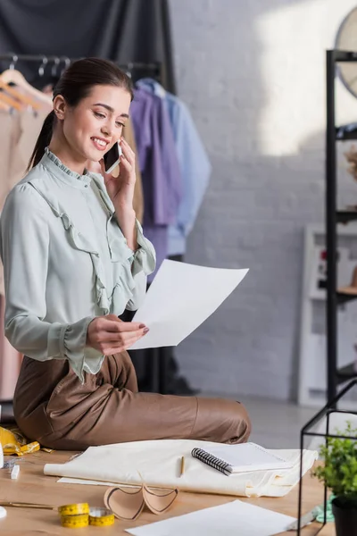 Positiver Designer spricht auf Smartphone in Tuchnähe, Nähmuster und verschwommenes Maßband — Stockfoto