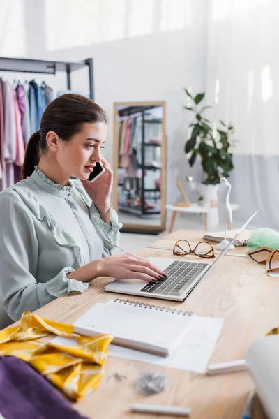 Seamstress talking on smartphone and using laptop near blurred cloth — Stock Photo