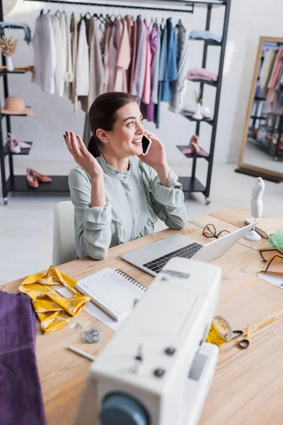 Smiling designer talking on smartphone near laptop and blurred sewing machine in atelier — Stock Photo