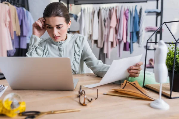 Designer with laptop holding paper near blurred tape measure and scissors — Stock Photo