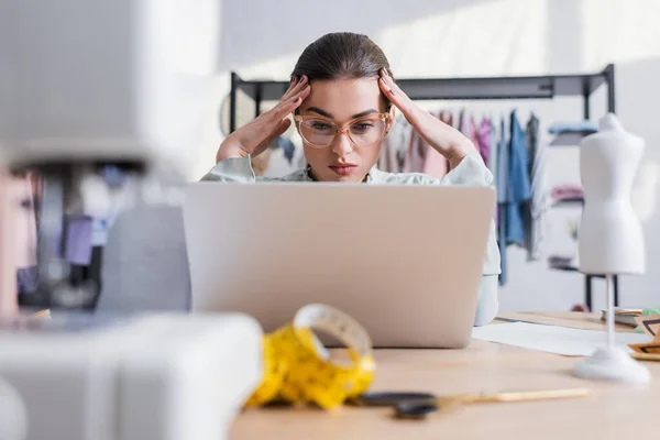 Designer in eyeglasses using laptop near blurred sewing machine and tape measure — Stock Photo