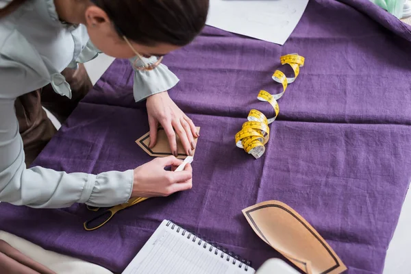 Overhead view of seamstress marking cloth and holding sewing pattern near scissors — Stock Photo