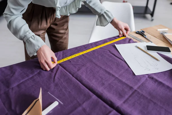 Vue recadrée du tissu de mesure de couturière près du téléphone cellulaire et croquis — Photo de stock