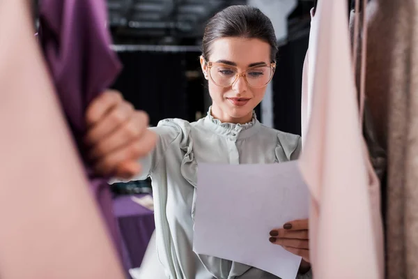 Seamstress in eyeglasses holding paper near blurred clothes — Stock Photo