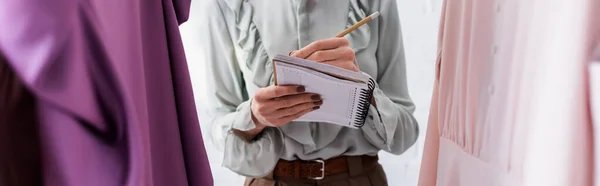 Vista recortada de la escritura del diseñador en el cuaderno cerca de la ropa en atelier, bandera - foto de stock