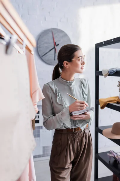 Side view of smiling seamstress writing on notebook near clothes and sewing pattern — Stock Photo