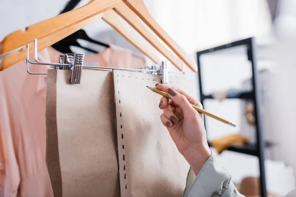 Vista recortada de costurera con lápiz cerca de patrones de costura en perchas — Stock Photo