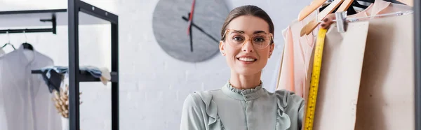 Smiling designer looking at camera near sewing patters on hangers, banner — Stock Photo