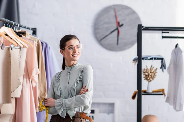 Diseñador sonriente con cinta métrica de pie cerca de patrones de costura y ropa en perchas - foto de stock