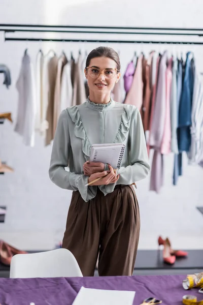 Cheerful seamstress holding notebook near blurred fabric — Stock Photo