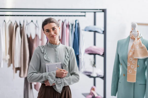 Designer with notebook looking at camera near mannequin with jacket — Stock Photo