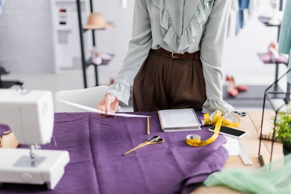 Cropped view of designer holding paper near smartphone, fabric and sewing machine — Stock Photo