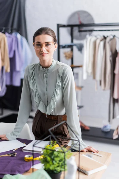 Smiling designer looking at camera near notebooks, fabric and tape measure — Stock Photo