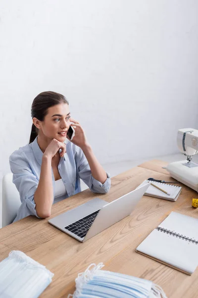 Sonriente costurera hablando en el teléfono inteligente cerca de la computadora portátil y máscaras médicas - foto de stock