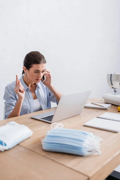 Cucitrice che parla su smartphone vicino a laptop, macchina da cucire e maschere mediche — Foto stock