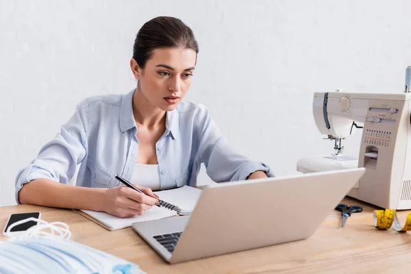 Seamstress writing on notebook near blurred laptop and medical masks — Stock Photo