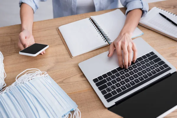 Cropped view of seamstress using smartphone and laptop near medical masks and notebooks — Stock Photo