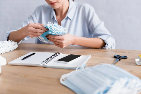 Cropped view of seamstress holding medical masks near smartphone and notebook — Stock Photo